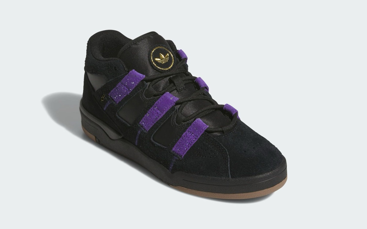 adidas Kader negro visto de frente en diagonal sobre fondo gris claro, con lengüeta de nylon negro y un parche redondo con el Trefoil dorado, cordones negros que pasan por las tiras de gamuza violeta moteada, puntera perforada y suela de goma color caramelo