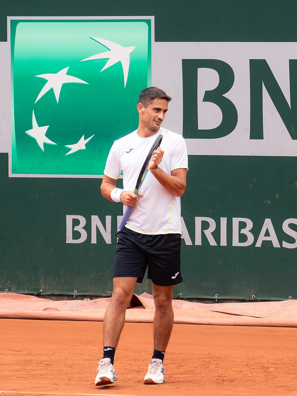 Foto de archivo: Mariano Navone, con remera blanca y short negro, camina por el polvo de ladrillo de Roland Garros con la raqueta en la mano, frente a un cartel verde de un sponsor del torneo