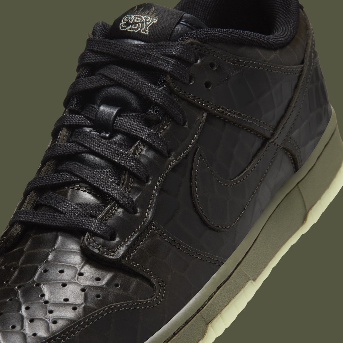Primer plano del Nike Dunk Low Shibuya Halloween con cordones negros, cuero negro texturado tipo cocodrilo y la sigla SBY bordada en la etiqueta de la lengüeta