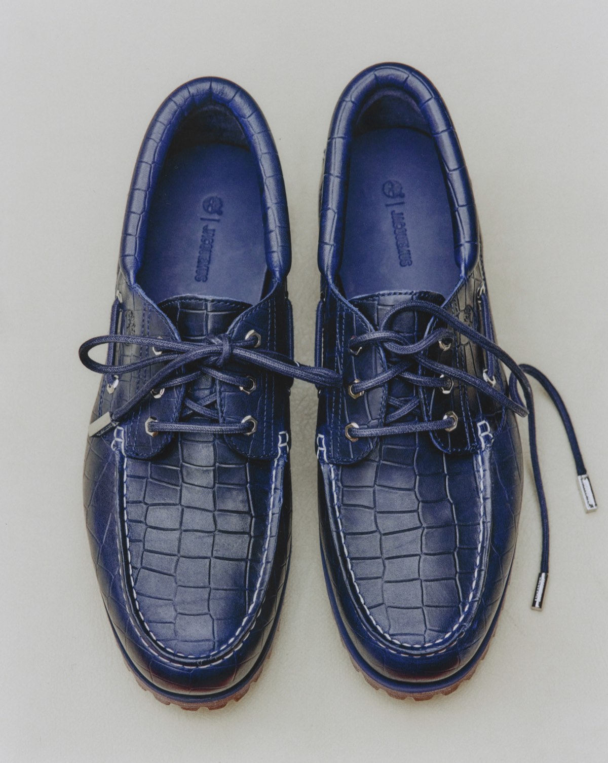 Par del Timberland x Jacquemus Authentic Boat Shoe en navy visto desde arriba sobre fondo gris claro, con todo el cuero en relieve de cocodrilo azul, costuras blancas en la puntera, ojales y herretes plateados, cordones azules y la plantilla azul con el árbol de Timberland y la palabra Jacquemus grabados