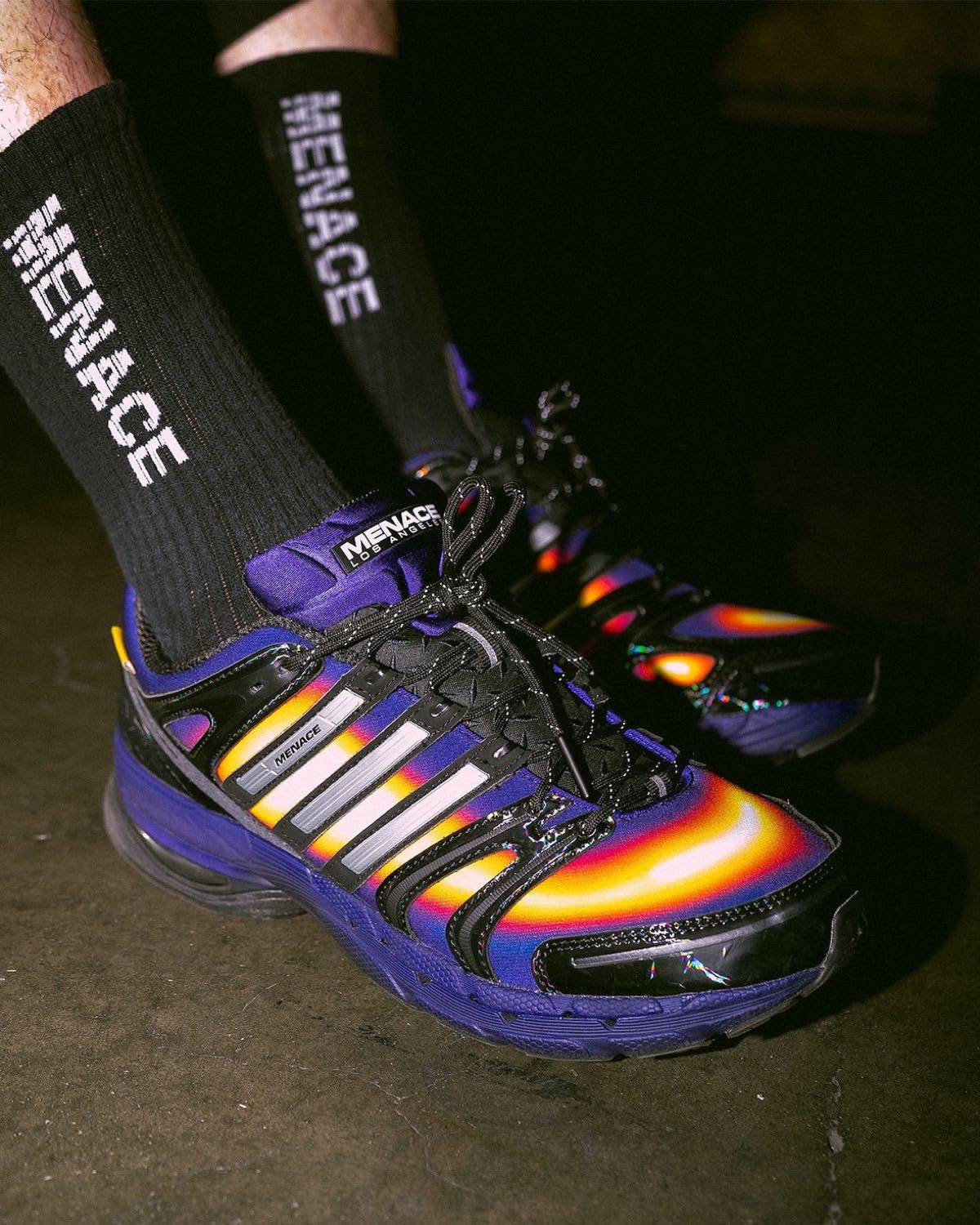 MENACE x adidas Adistar Control 5 Heat Signature puesto, de perfil lateral, con el degradé amarillo y rojo asomando entre los overlays negros, Tres Tiras plateadas con la palabra MENACE, etiqueta MENACE Los Angeles en la lengüeta y medias negras con el logo de la marca