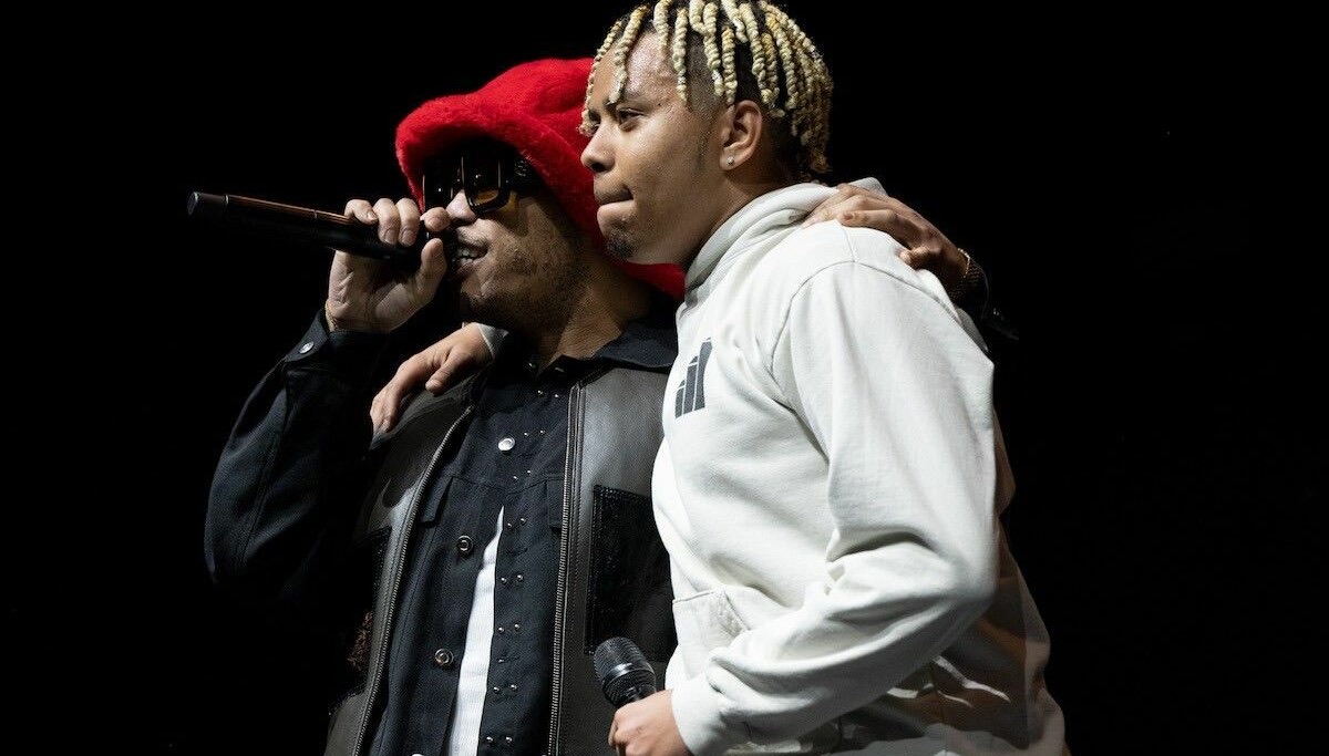 Anderson .Paak, con gorro rojo de peluche y anteojos oscuros, canta con un micrófono mientras abraza a Cordae, de buzo gris claro y trenzas rubias, sobre un escenario con fondo negro