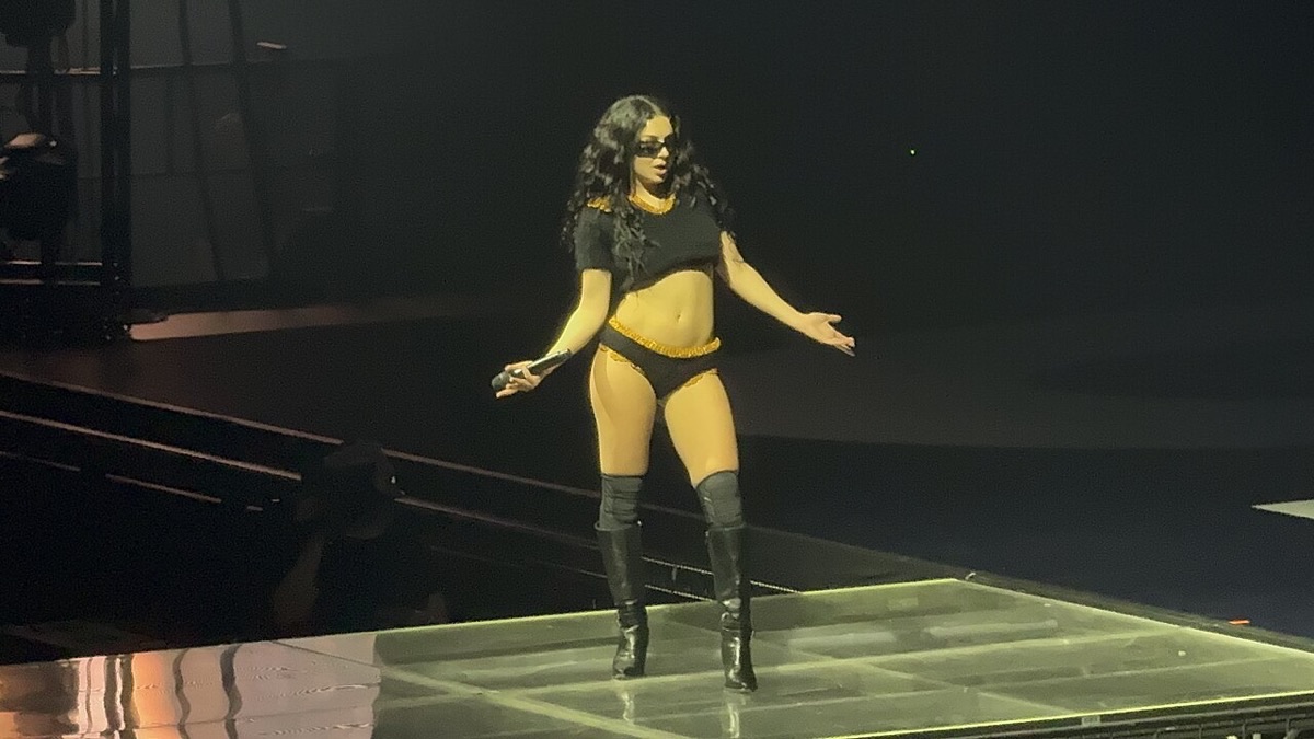 Foto de archivo de Charli XCX en el escenario en abril de 2025: con anteojos de sol, top negro corto, bombacha amarilla y botas negras altas, abre los brazos con el micrófono en la mano sobre una plataforma iluminada. Foto: Erin Mc, Wikimedia Commons, CC BY 2.0