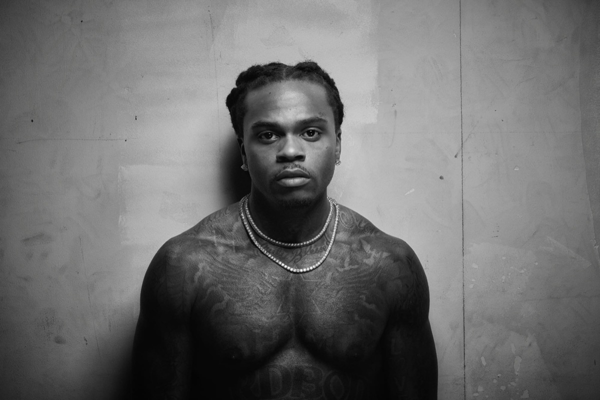 Foto de prensa oficial de Gunna para "Pop It", en blanco y negro: el rapero con el torso tatuado, dos cadenas plateadas y trenzas, mirando a cámara contra una pared de cemento