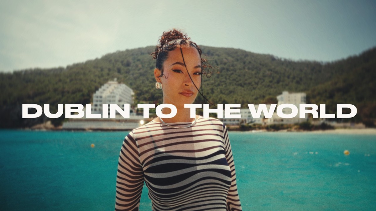 Fotograma del mini documental oficial "Dublin to the World": Jazzy con remera rayada blanca y negra, frente a un mar turquesa y una costa con hoteles blancos, con el título del film sobreimpreso