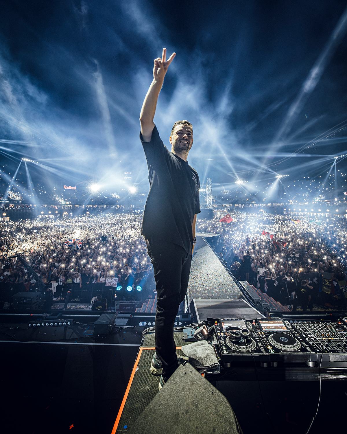 Martin Garrix de espaldas al público en un festival, con la mano en alto haciendo el signo de la paz, sobre la cabina de DJ, frente a una multitud iluminada por luces azules (foto de archivo)