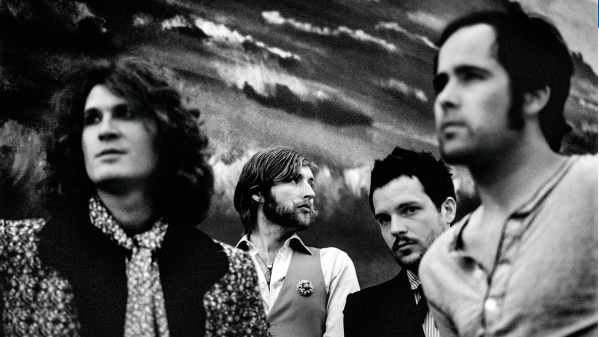 Foto de archivo en blanco y negro de The Killers en la época de Sam's Town: los cuatro integrantes delante de un fondo pintado con un cielo nublado, Brandon Flowers de saco oscuro mirando a cámara