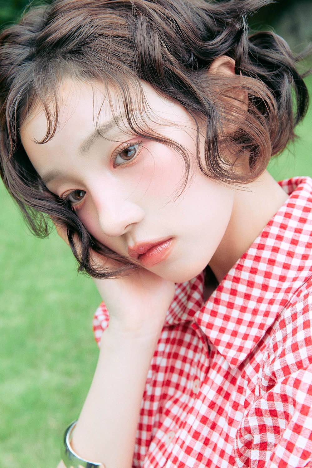 Retrato de prensa de YUQI con el pelo corto y ondulado, camisa a cuadros rojos y blancos y la mano apoyada en la mejilla, sobre un fondo de pasto. Foto: Cube Entertainment