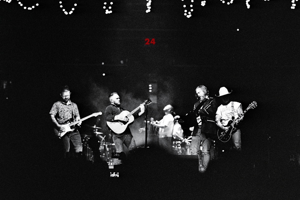 Imagen oficial de archivo de la web de Zach Bryan: foto en blanco y negro de Bryan y su banda en vivo, con guitarras eléctricas y acústica, bajo guirnaldas de luces y un 24 en rojo al fondo del escenario