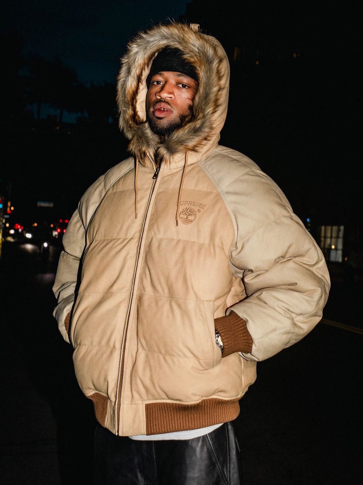 Modelo de noche en la calle con la campera Supreme x Timberland Suede Puffer color wheat, con capucha de piel sintética y gorro oscuro