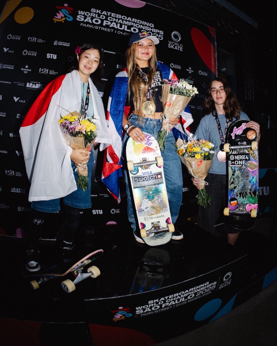 Sky Brown, envuelta en la bandera británica, sonríe con la medalla de oro, flores y su tabla en el podio del Mundial de San Pablo, con Mizuho Hasegawa a su derecha