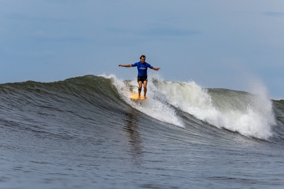 Tony Silvagni, con lycra azul, camina sobre un longboard amarillo con los brazos abiertos en una ola de El Sunzal