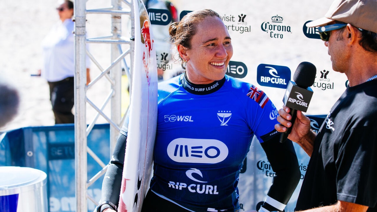 Carissa Moore, con la lycra azul del MEO Rip Curl Pro Portugal y la bandera de Hawái, sonríe con su tabla mientras la entrevistan frente al panel del evento en Supertubos