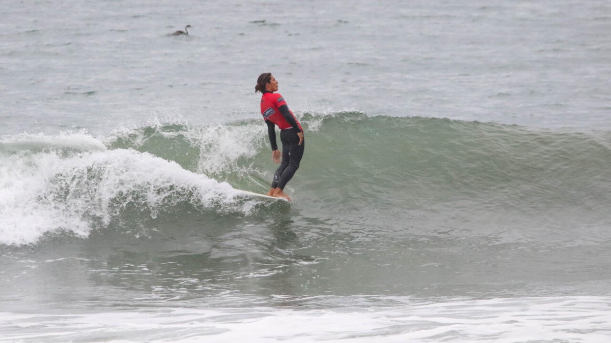 Jonathan Melendres, con lycra roja y traje negro, se inclina hacia atrás parado sobre su longboard mientras la ola se cierra a su espalda en Pismo Beach