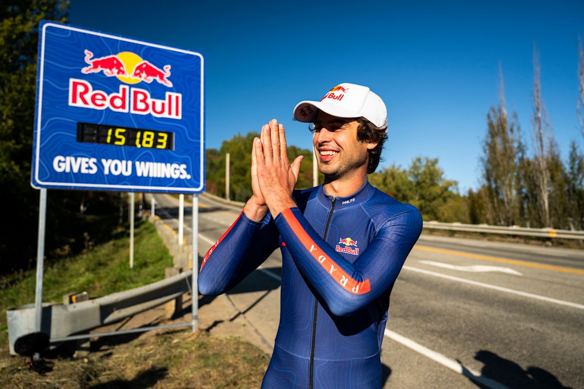 Diego Poncelet, con gorra blanca y traje aerodinámico azul, junta las manos y sonríe al costado de la ruta, junto a un cartel que marca 151.83