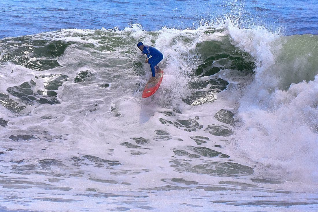 Un surfista con traje azul y tabla roja baja una ola grande que rompe en espuma en Punta Rocas, Perú