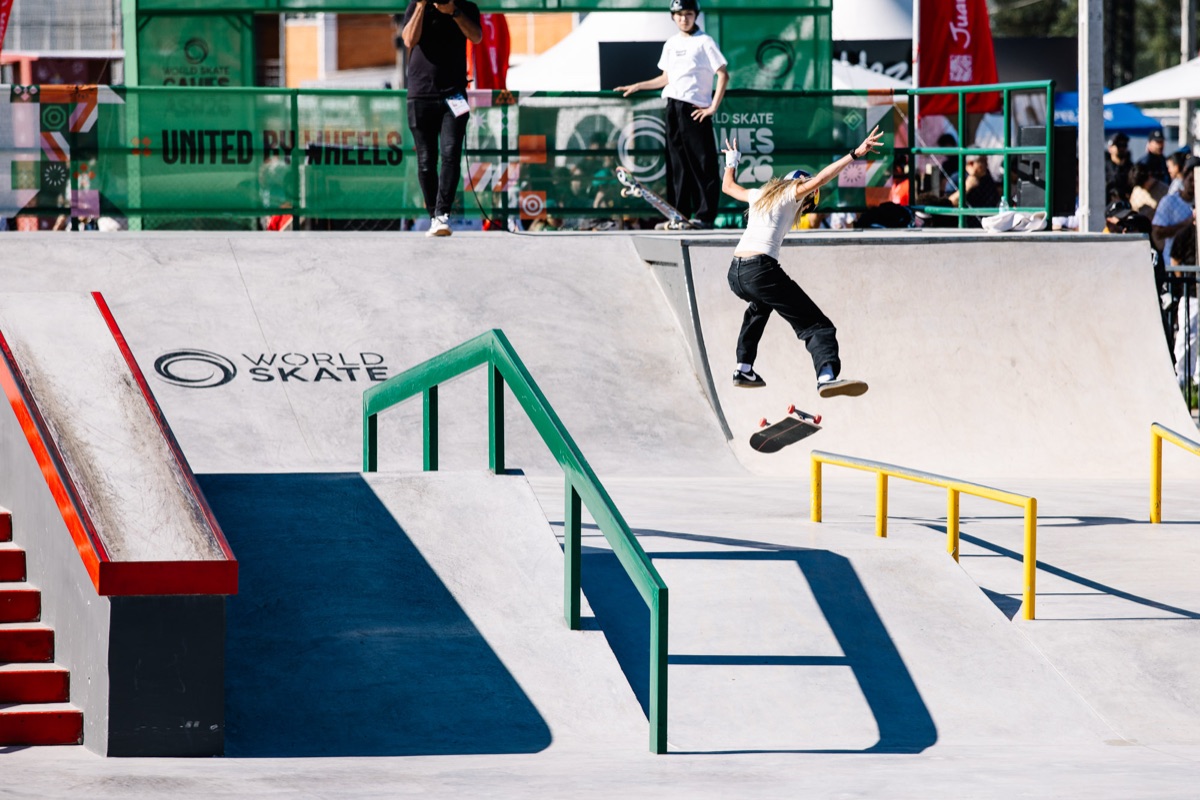 Chloe Covell, con remera blanca y pantalón negro, salta en el aire con la tabla girando debajo, junto a la baranda verde de la pista de street de Luque