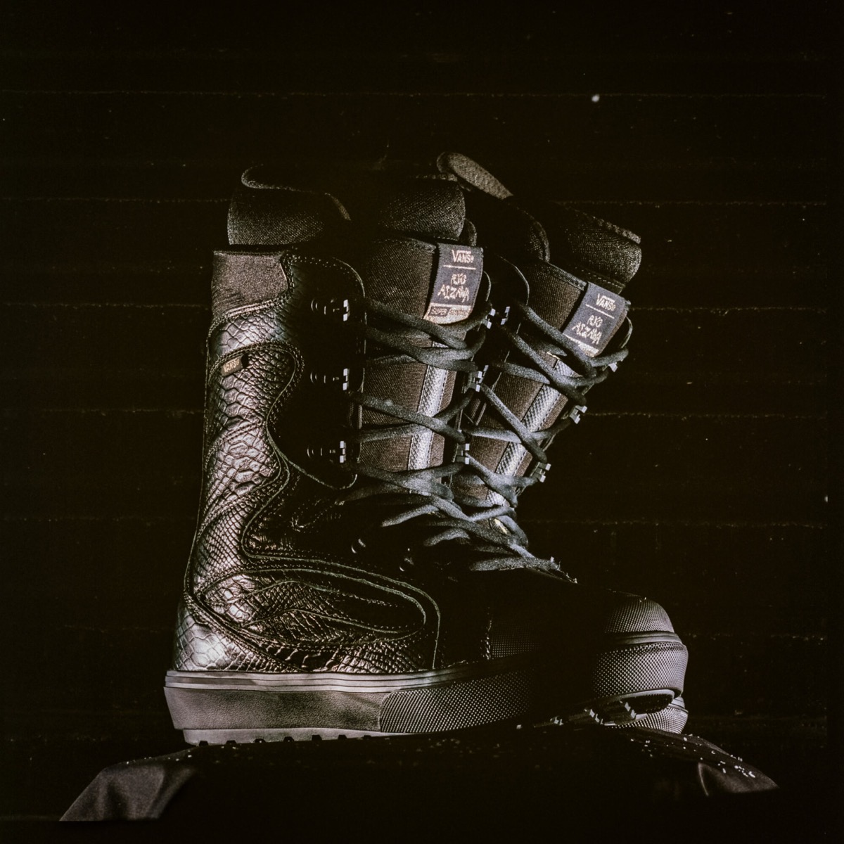 Par de botas de snowboard Vans Super Standard negras con textura de serpiente, cordones negros y lengüeta con la firma de Ryo Aizawa, fotografiadas sobre fondo oscuro