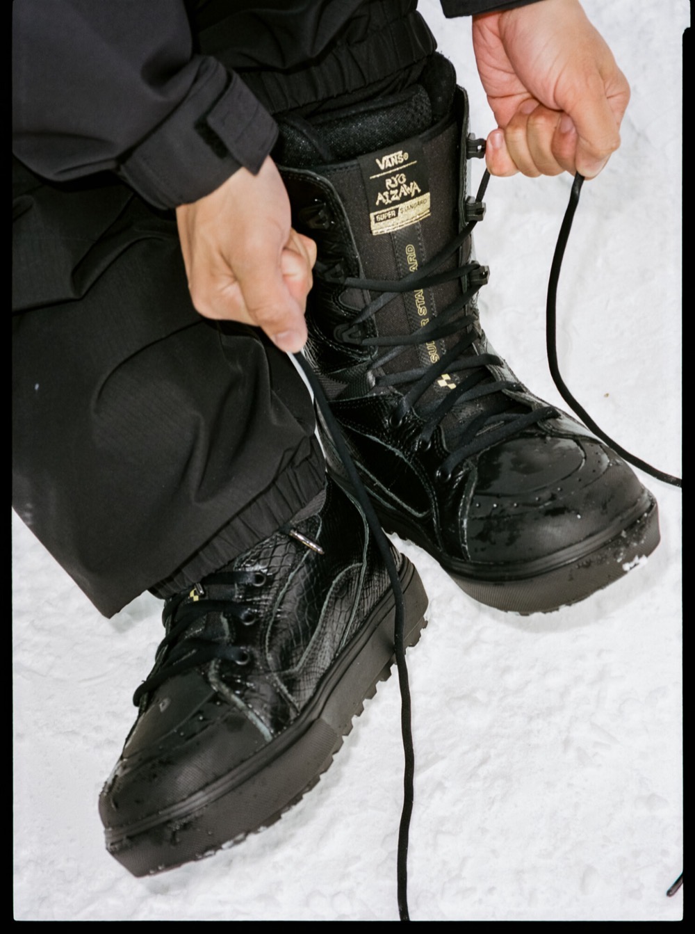 Rider atándose los cordones de las botas Vans Super Standard negras con textura de serpiente sobre la nieve; en la lengüeta se lee la firma de Ryo Aizawa